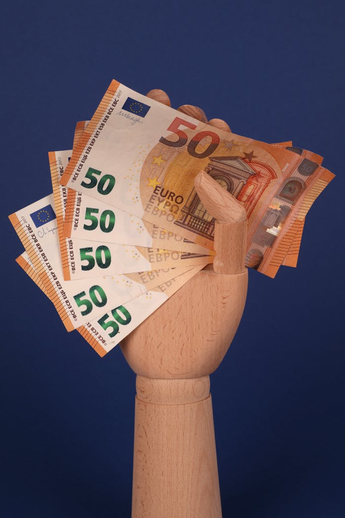 Wooden hand holding multiple euro banknotes against a blue backdrop, symbolizing finance and wealth.